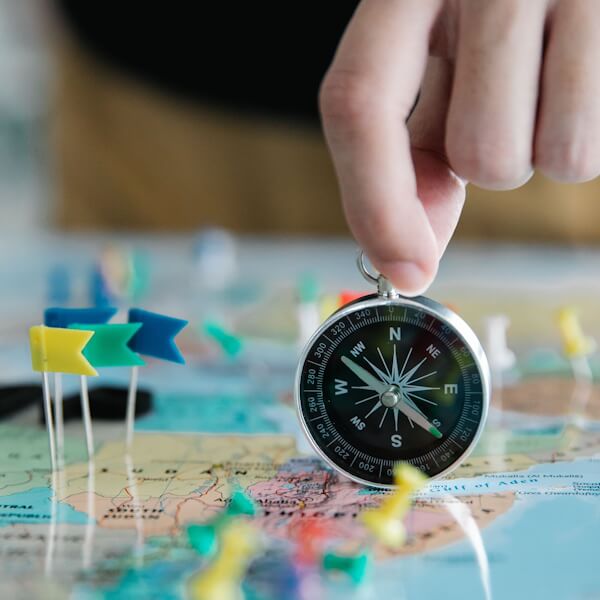 A back-and-white compass is held by by a hand on a colorful map with pushpins, photograph by Lara Jameson of Pexels